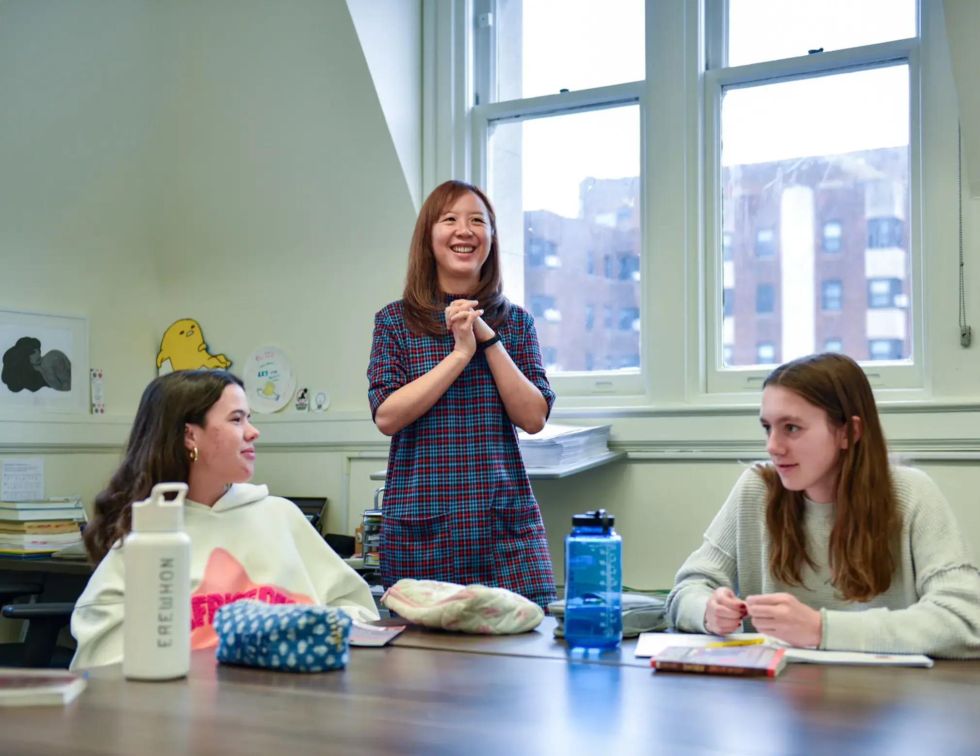 three girls in classroom, two sitting and the one in the middle standing with her hands crossed and smiling